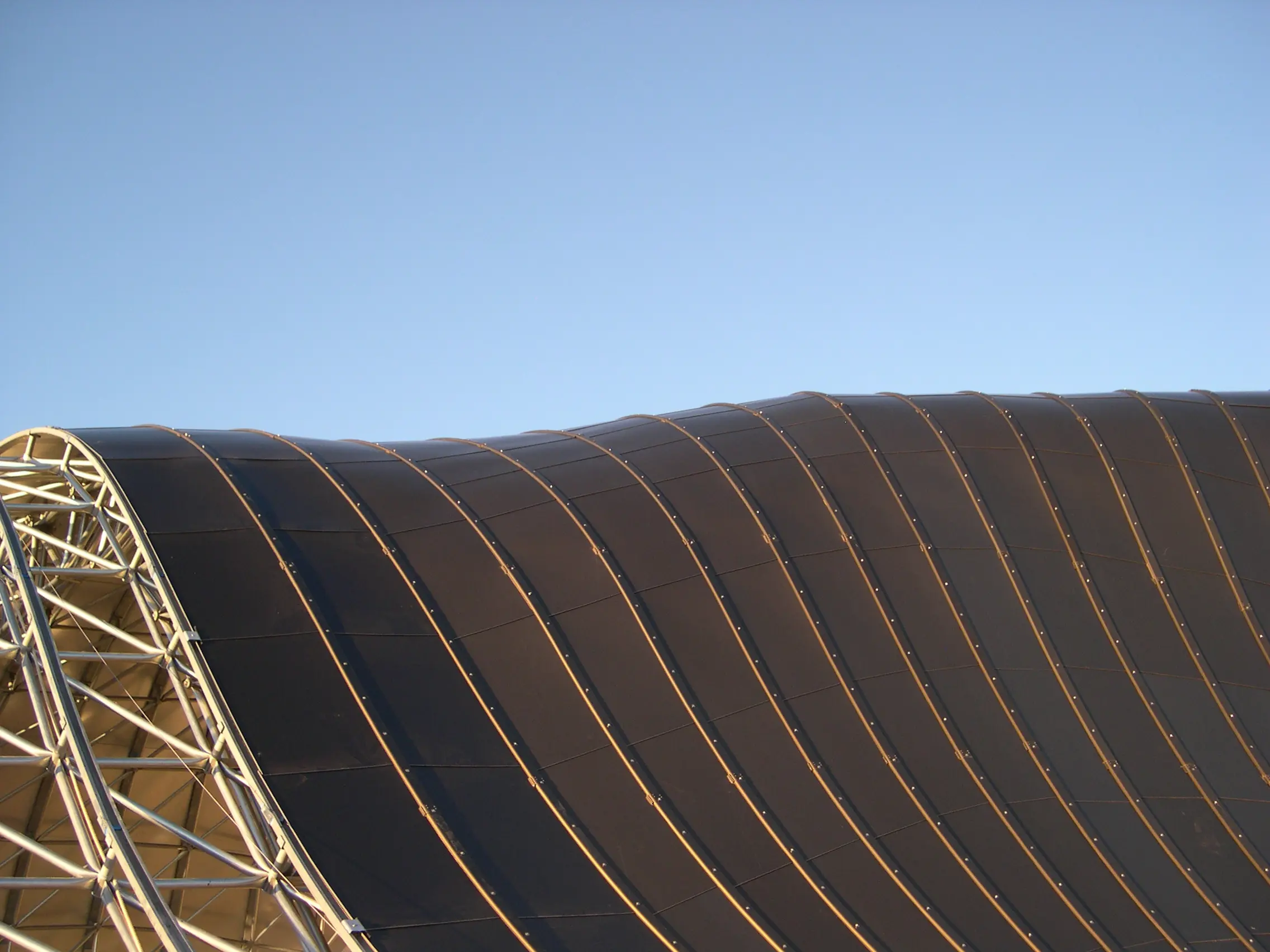 Revestimiento metálico en techo tipo bóveda de cañón con paneles curvos y borde de estructura space frame visible, mostrando disposición de paneles, fijaciones e integración estructural en cubiertas de gran luz.