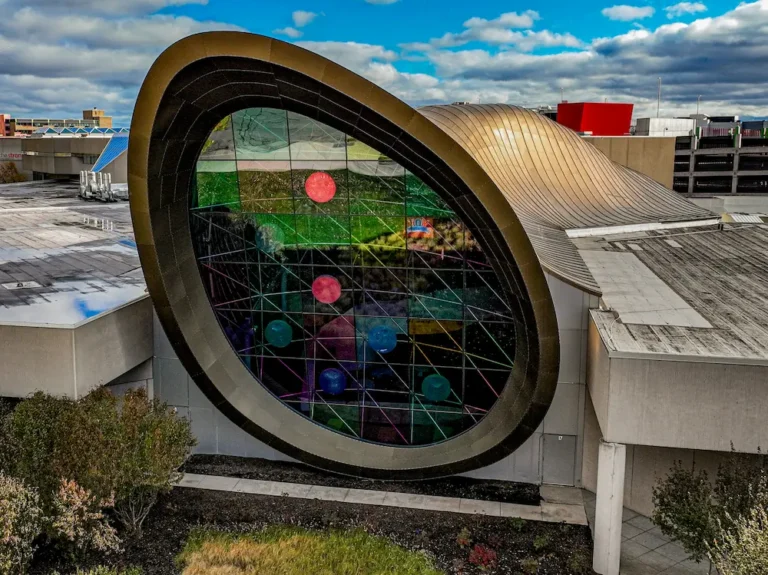 Structure architecturale de forme libre avec toit métallique courbé et grande façade ovale en verre avec motifs géométriques et éléments colorés, intégrée dans un environnement urbain moderne.