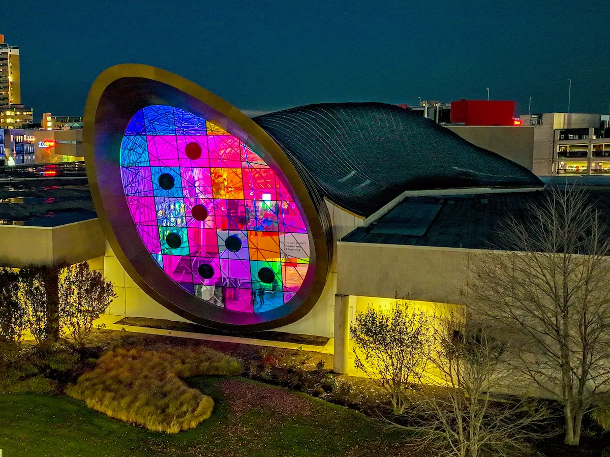 Edificio de forma libre tipo bóveda durante la noche con fachada de vidrio iluminada y estructura space frame visible, destacando iluminación arquitectónica, integración de color y diseño urbano.
