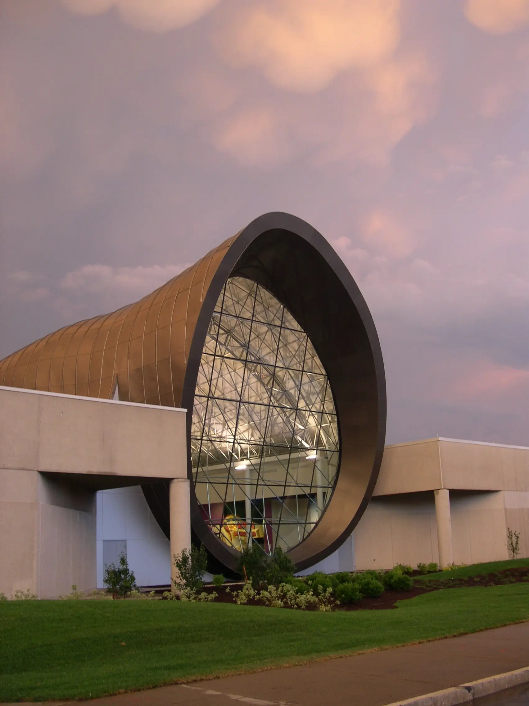 Vista exterior de una estructura tipo bóveda de forma libre con gran fachada de vidrio y sistema space frame visible, integrada en un diseño arquitectónico moderno.