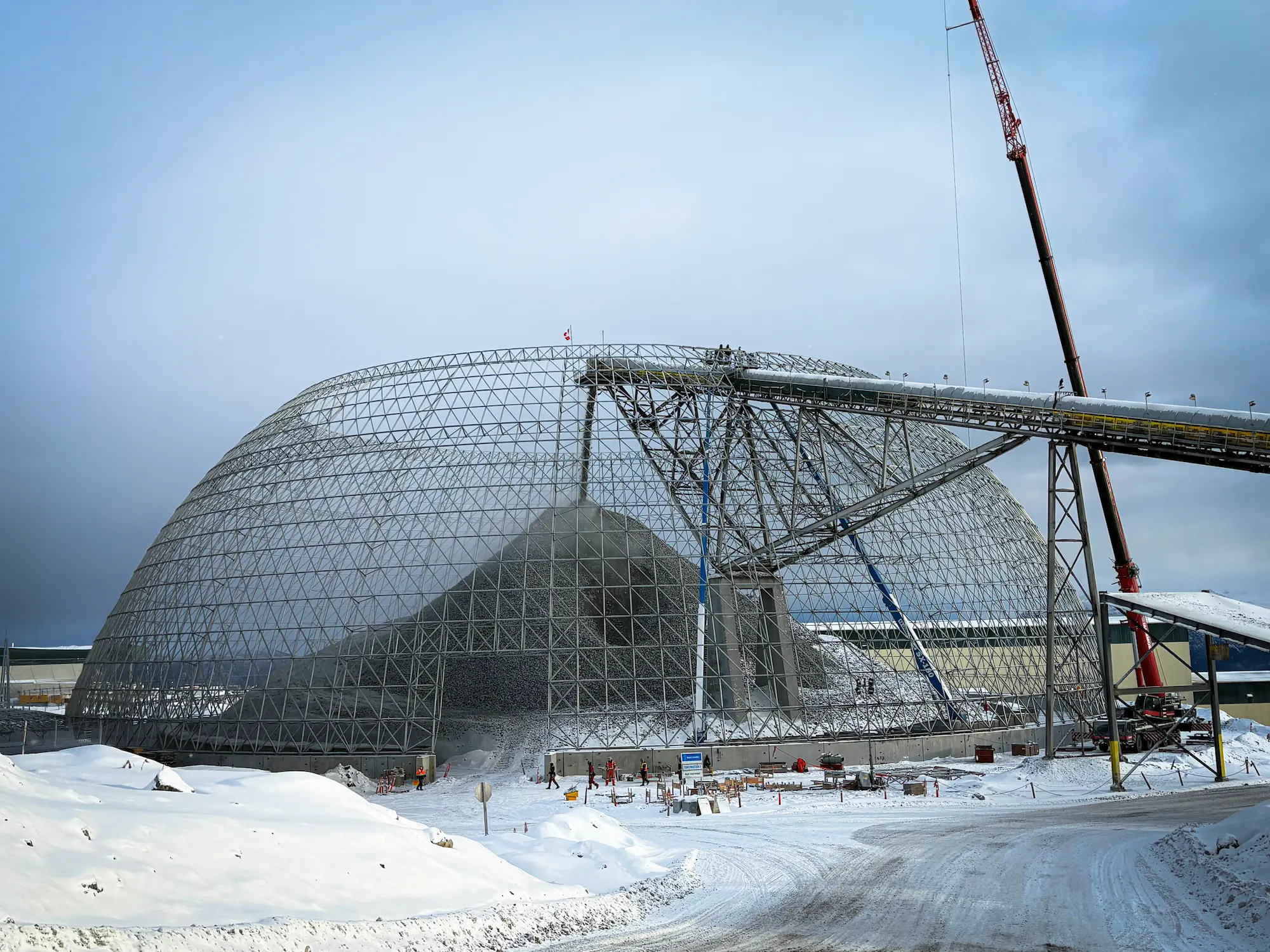 Triodetic core geodesic dome structure under construction at a snowy industrial site with a crane and elevated conveyor.