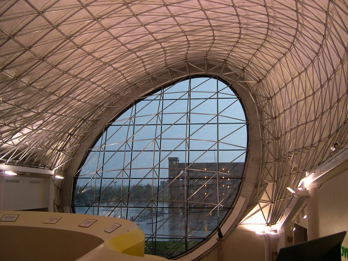 Interior de una estructura space frame tipo bóveda de cañón con gran fachada circular de vidrio, mostrando entramado estructural, entrada de luz natural y detalles arquitectónicos en un espacio público.