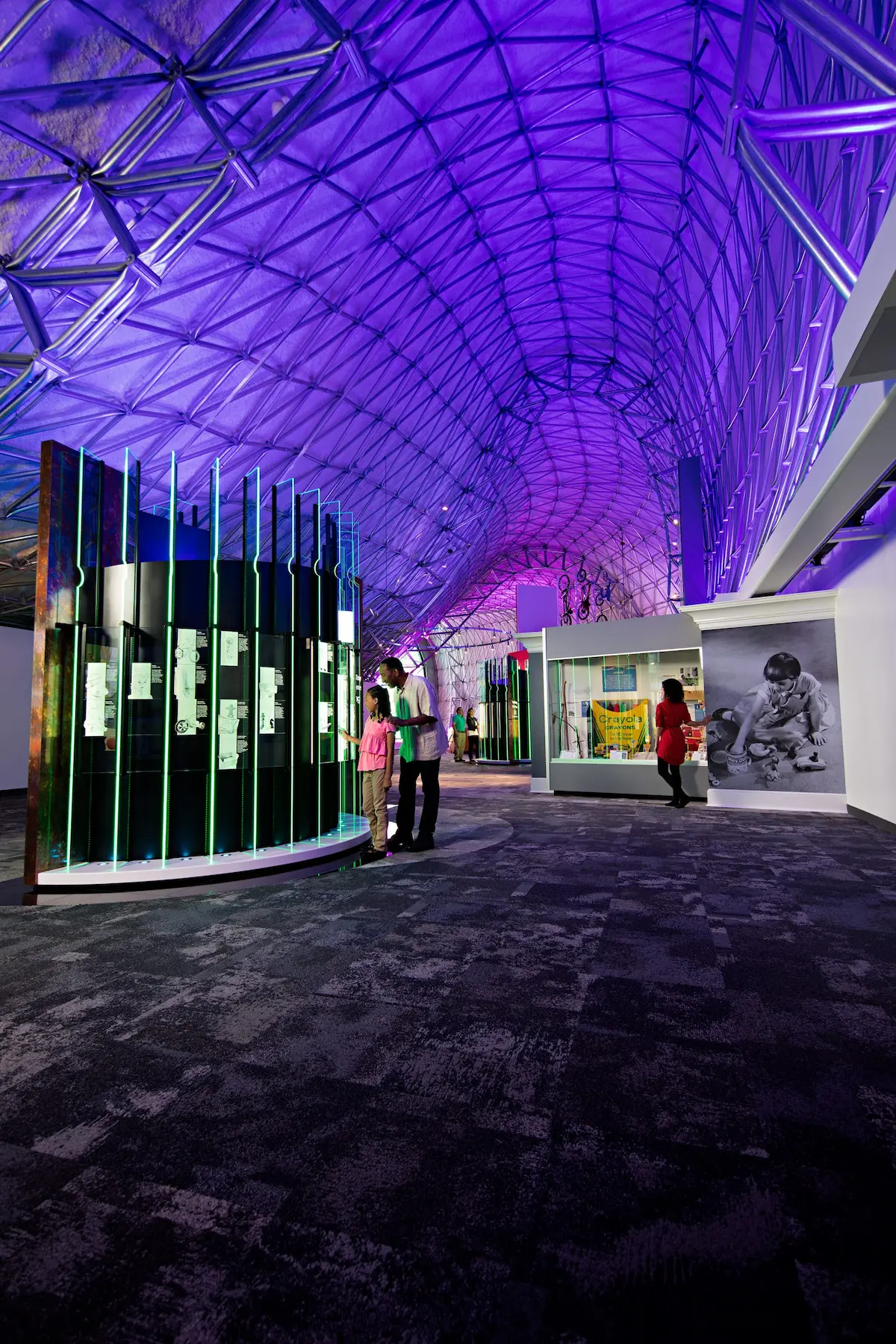 Vista interior de una estructura space frame con techo curvo de acero y sistema geométrico reticulado, utilizada en una sala de exhibición moderna con iluminación y elementos interactivos.